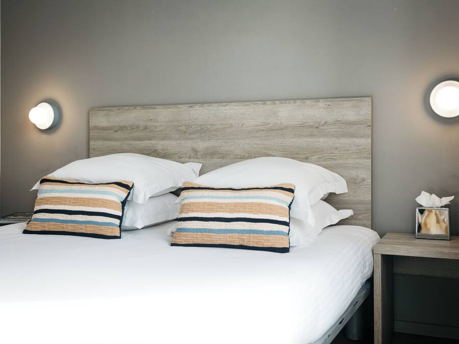 Una cama bien hecha con sábanas blancas, dos almohadas a rayas, un cabecero de madera y lámparas de noche.