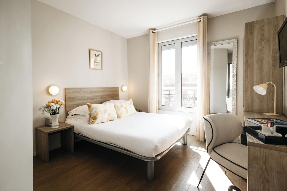 Quarto de hotel moderno com cama, mesas de cabeceira, escrivaninha, cadeira, janela com cortinas e espelho fixado na parede.
