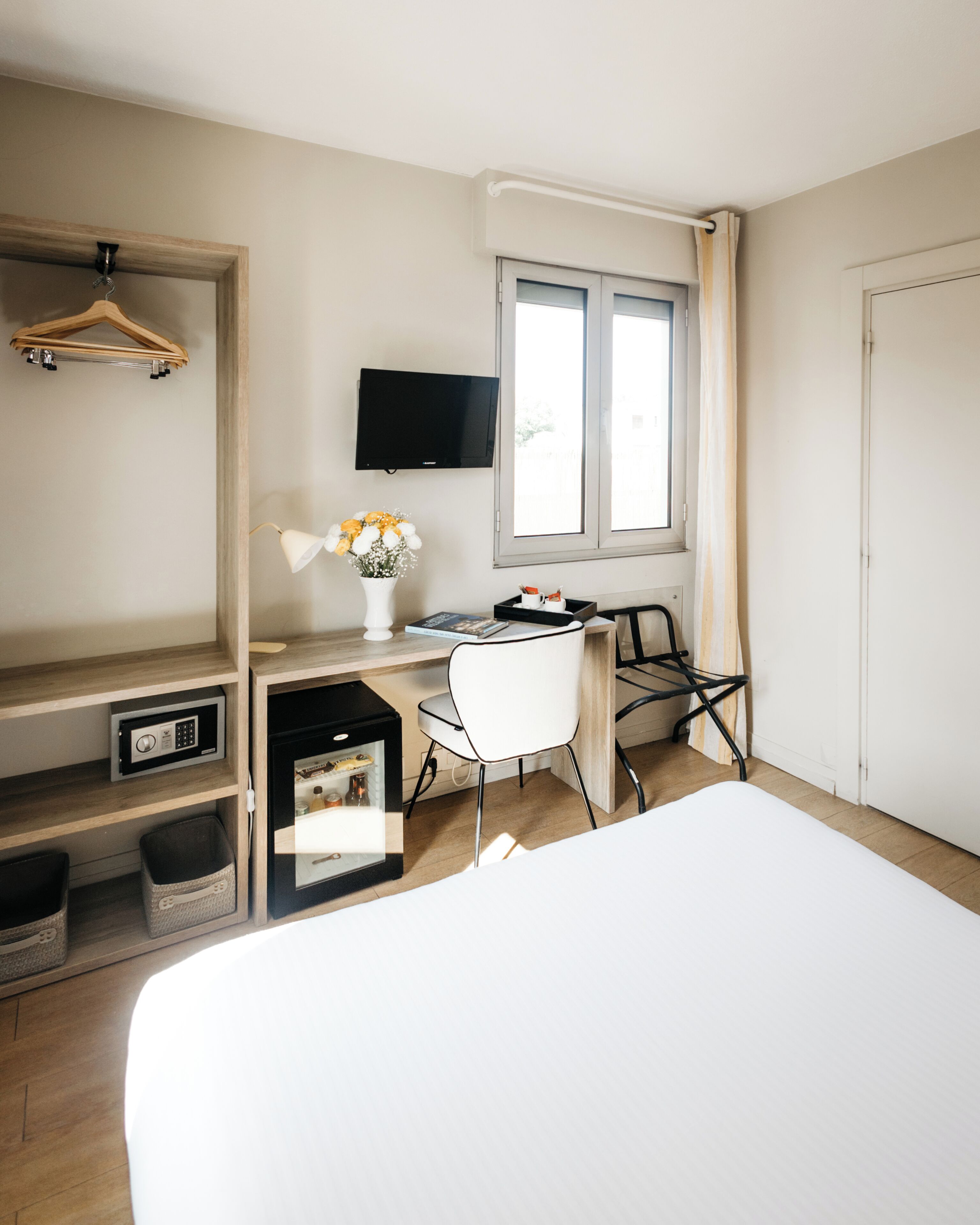 A hotel room with a bed, a desk with a chair, a television, a window with curtains, and a hanging clothes rack.