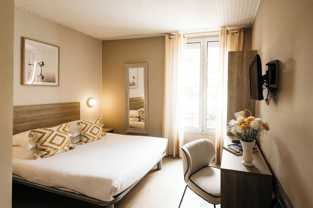 Quarto de hotel com cama, mesa de cabeceira, cadeira, escrivaninha, televisão e janela com cortinas.