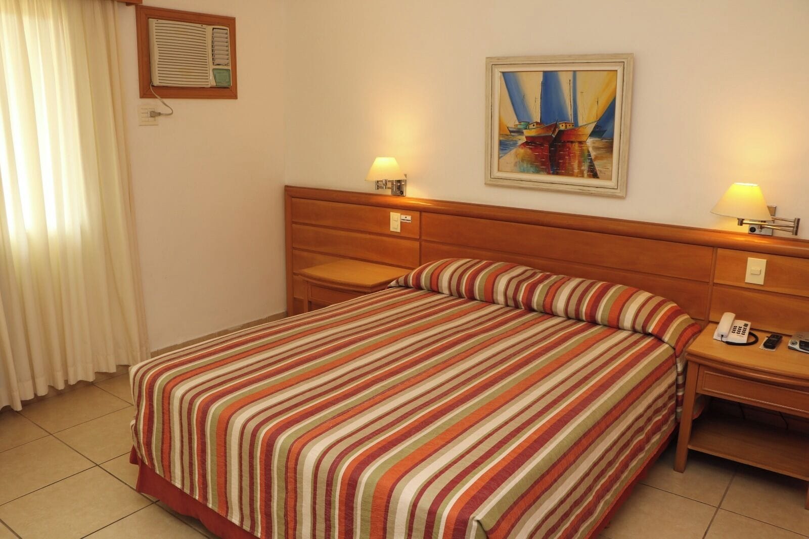 A hotel room with a bed, two bedside tables, a nightstand, a painting on the wall, and a window with curtains.