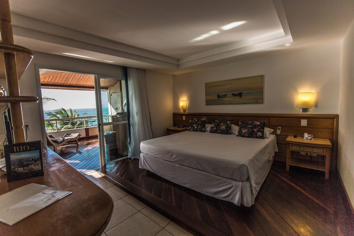 Hotelkamer met een bed, bureau, stoel, nachtkastje, lamp, schilderij en uitzicht op de oceaan.