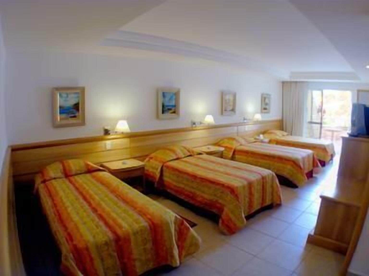 Habitación de hotel con tres camas, cada una con cubrecama a rayas, cabecero de madera y mesita de noche con lámpara. En la pared hay cuadros enmarcados y un televisor sobre un mueble.