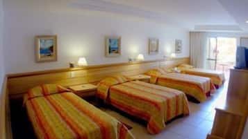 Habitación de hotel con tres camas, cada una con cubrecama a rayas, cabecero de madera y mesita de noche con lámpara. En la pared hay cuadros enmarcados y un televisor sobre un mueble.