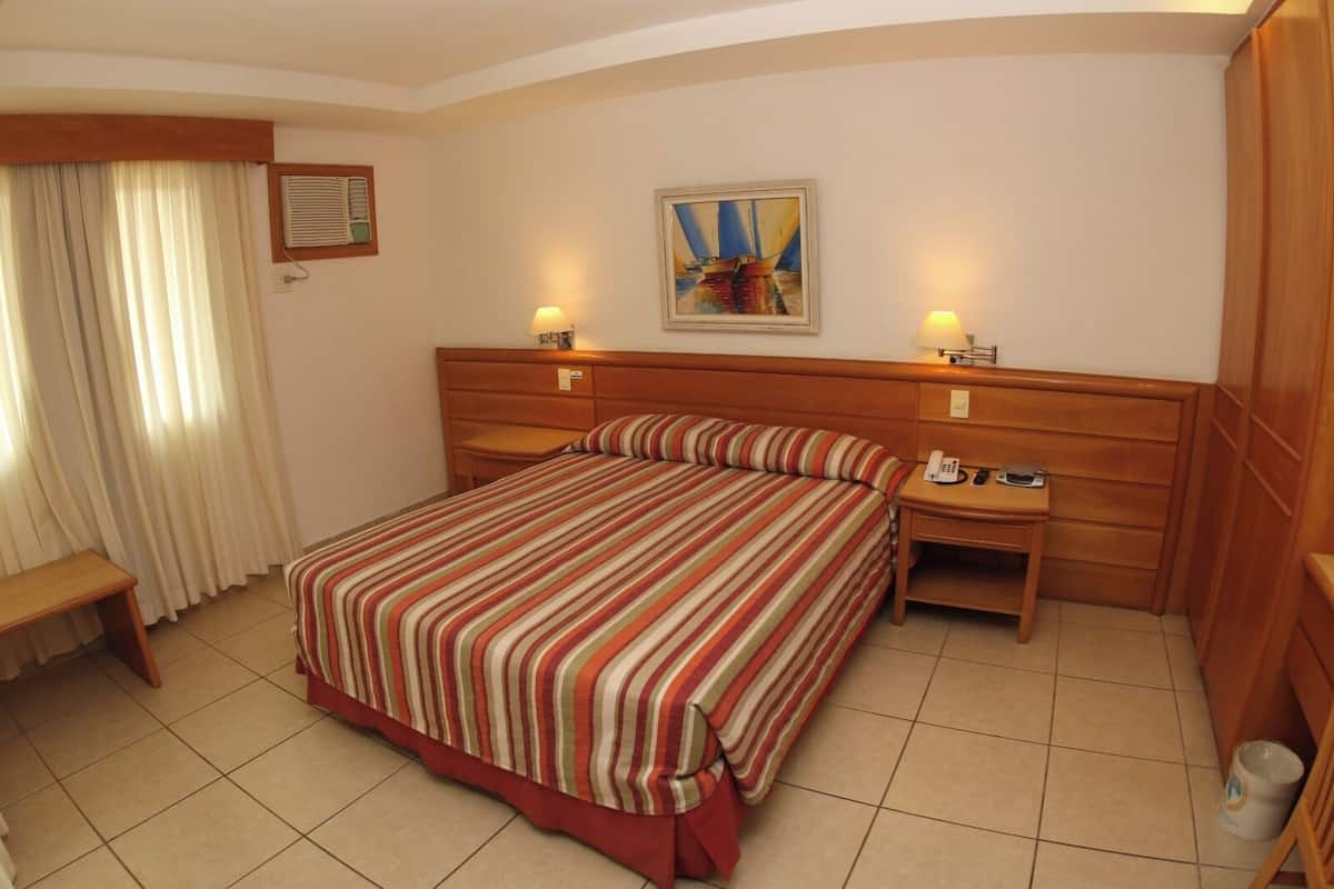 Een hotelkamer met een groot bed, twee nachtkastjes, een bureau, een stoel, een raam met gordijnen en een schilderij aan de muur.