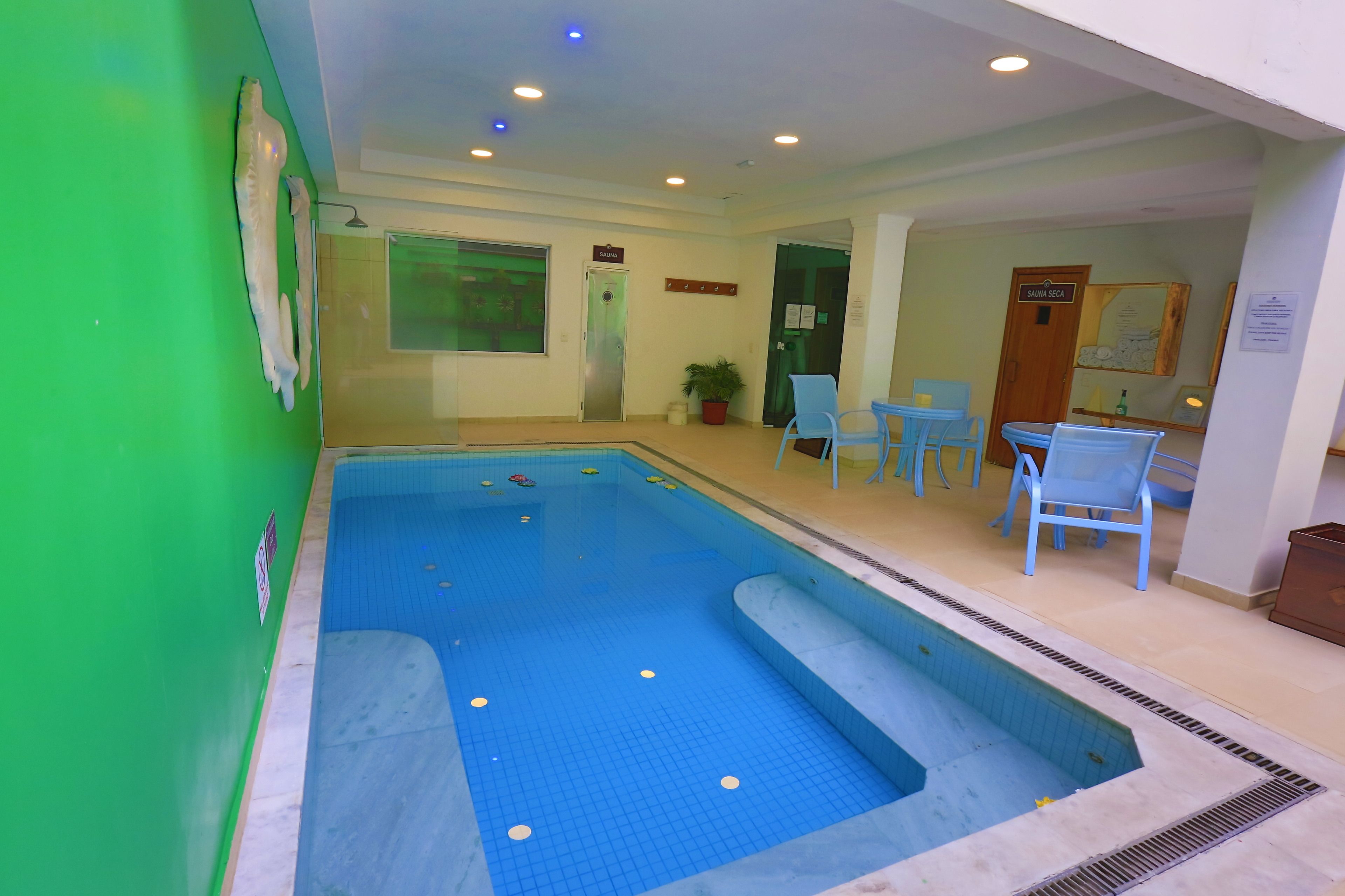 Una piscina interior con suelo de azulejos azules, una zona de estar con sillas azules y un cuarto de sauna.