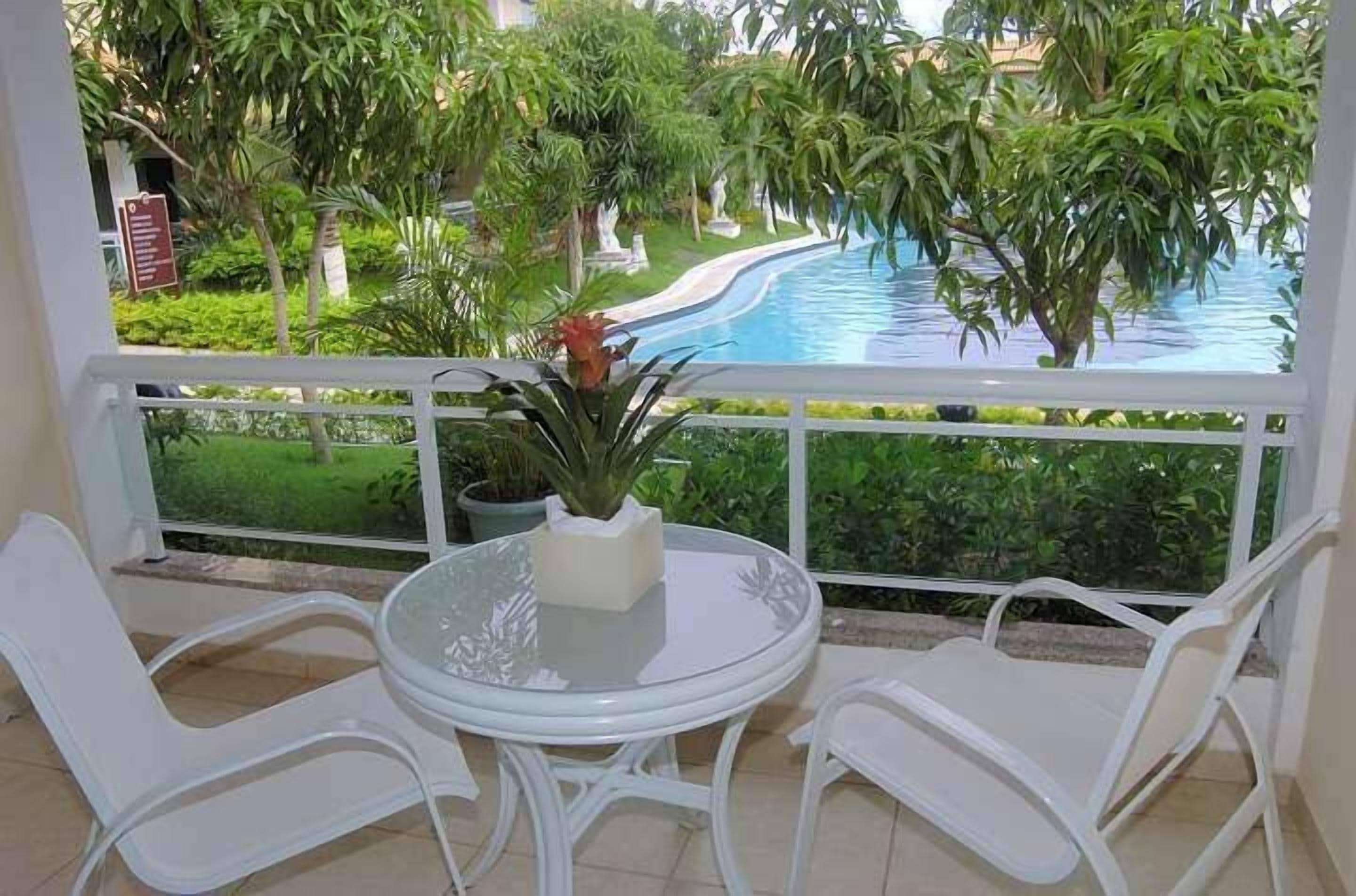 A balcony with a glass table and white chairs, overlooking a swimming pool and lush greenery.