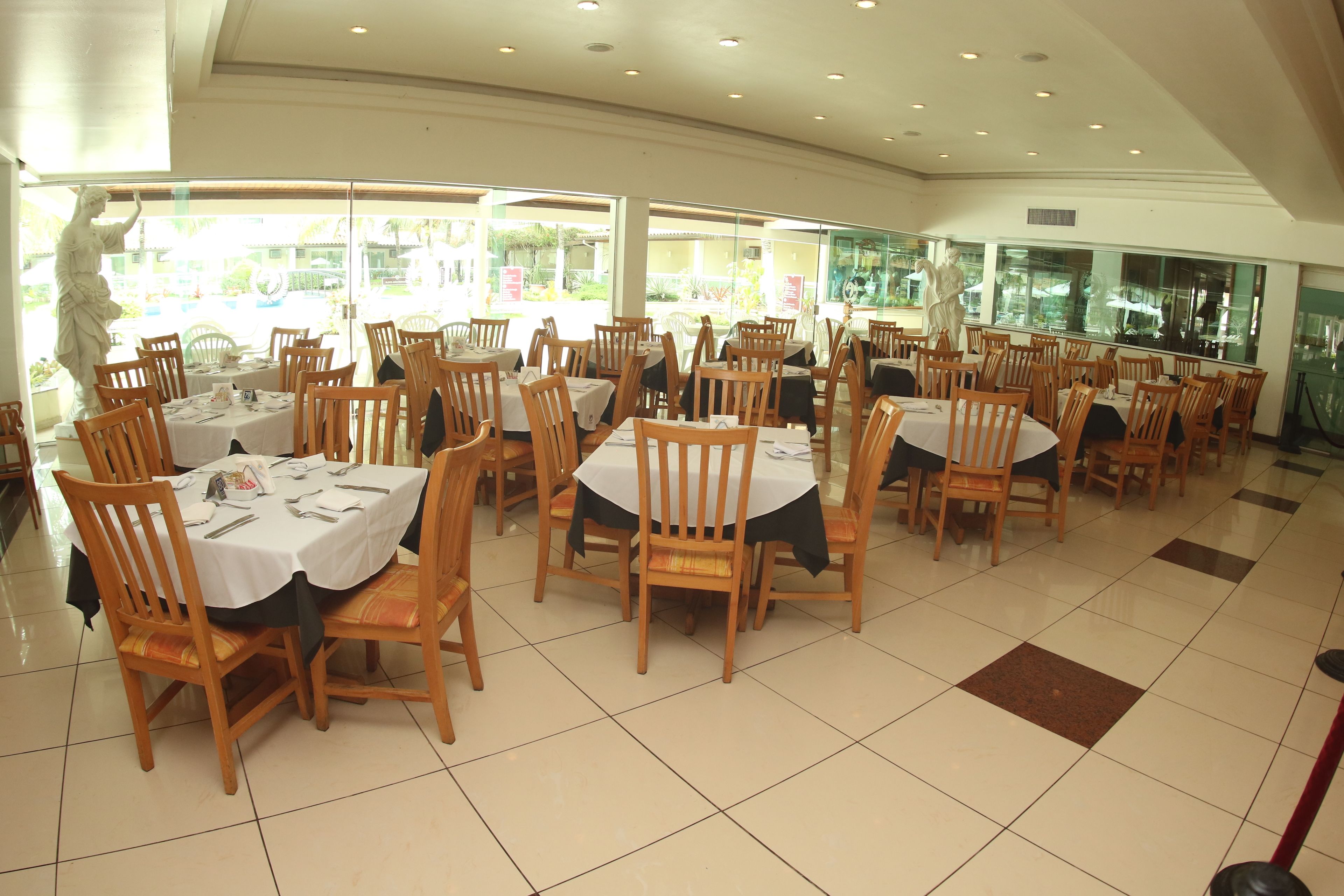 Un restaurante con mesas redondas y sillas de madera, grandes ventanales con vistas y suelo de baldosas.