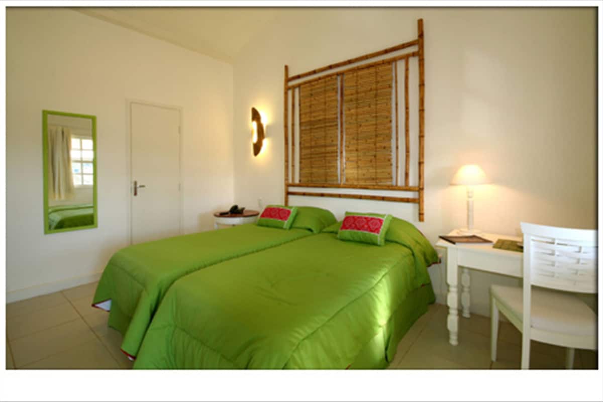 Ein Zimmer mit einem Bett, einem Schreibtisch, einem Stuhl, einer Lampe, einem Spiegel und einem Fenster mit Bambusrollvorhang.
