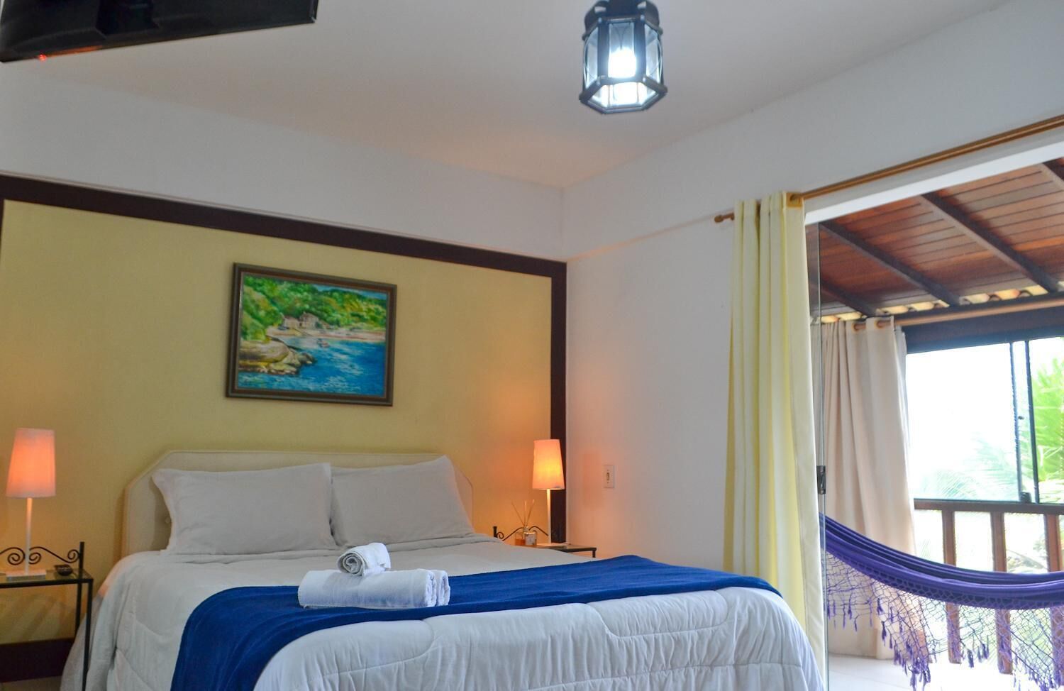 A hotel room with a bed, a painting, a lamp, and a view of the outdoors.