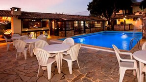 Un área de piscina al aire libre con suelo de piedra, sillas y mesas de plástico blanco.