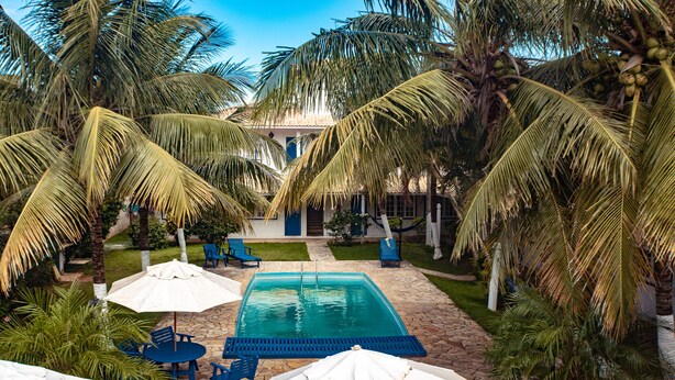 A pool area with blue chairs and umbrellas, surrounded by palm trees and a building with a blue door. - Pousada Luar de Buzios (Búzios)