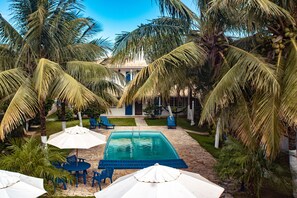 A pool area with blue chairs and umbrellas, surrounded by palm trees and a building with a blue door. - Pousada Luar de Buzios (Búzios)