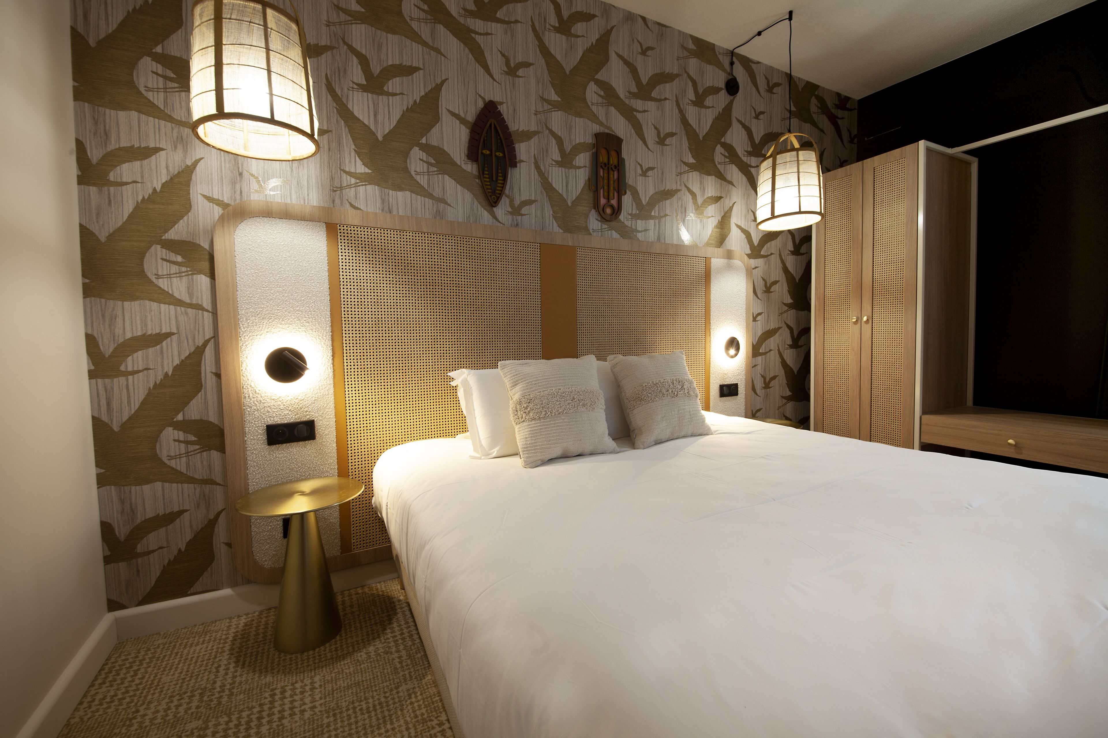 Ein modernes Hotelzimmer mit einem großen Bett, zwei Wandlampen, einem runden Beistelltisch und einem großen Kleiderschrank.
