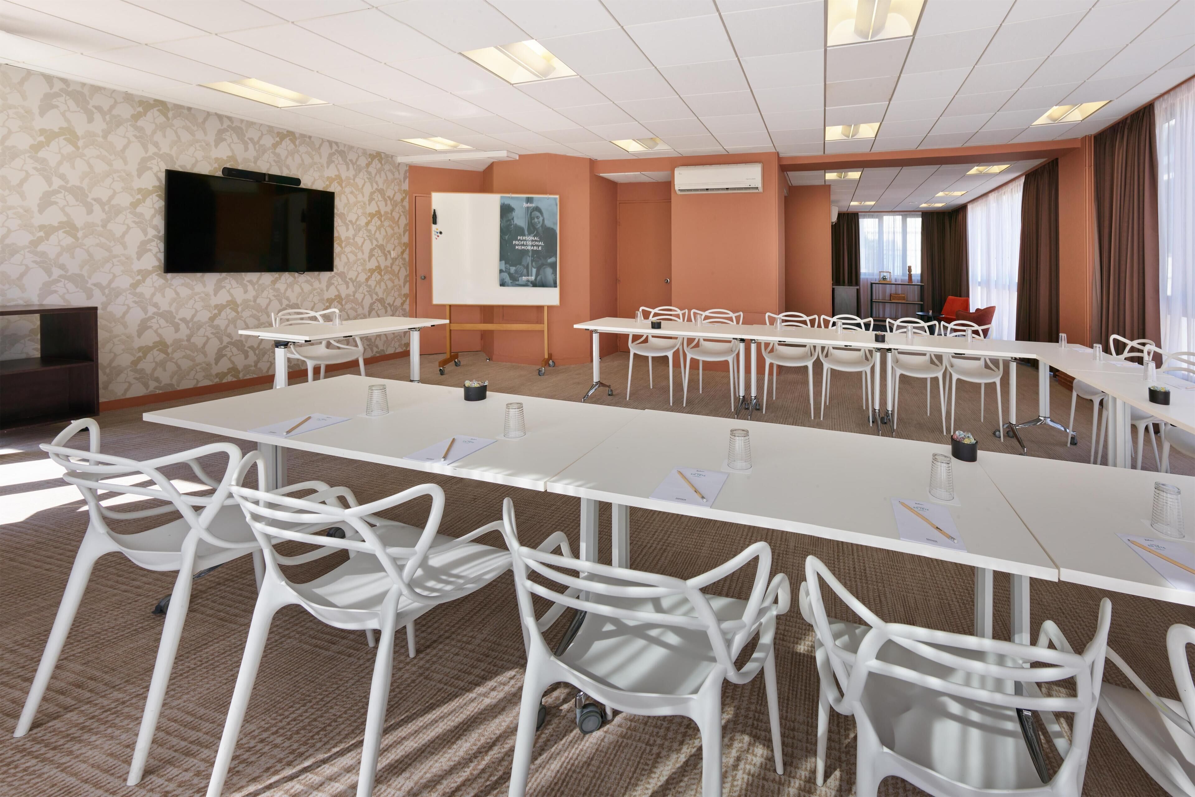 A conference room with a long table, white chairs, a flat-screen TV, a whiteboard, and a poster.
