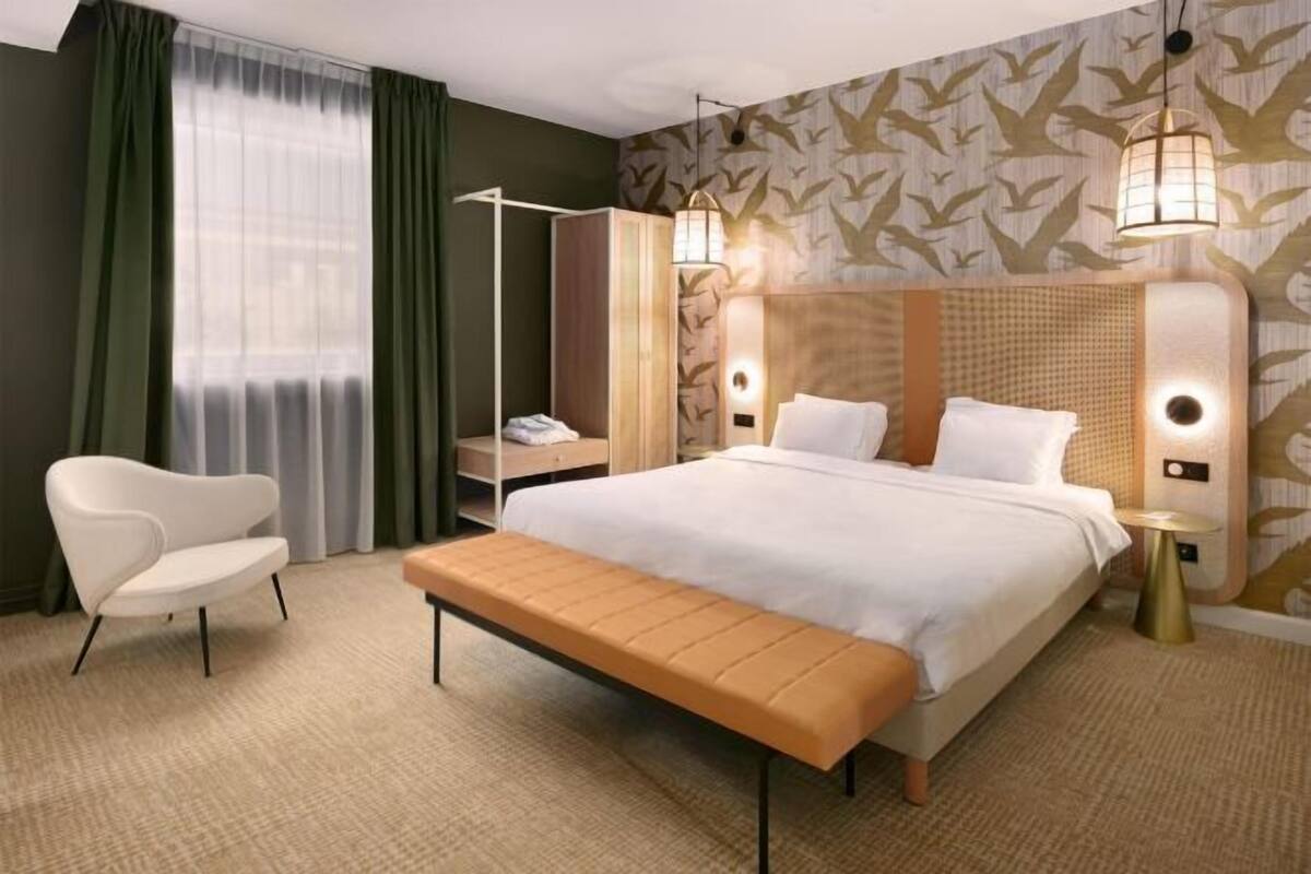 Habitación de hotel con una cama grande, una mesita de noche, una silla, un banco y una ventana con cortinas.