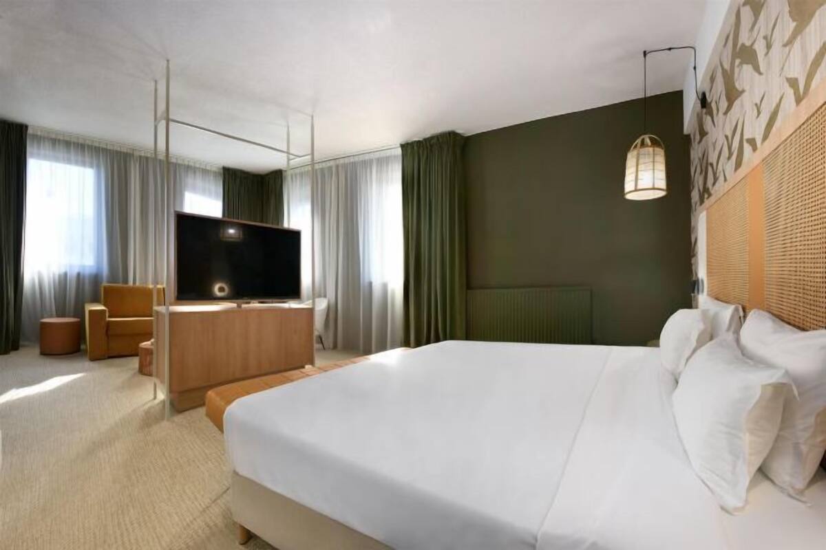 Una habitación de hotel con una cama grande, un televisor de pantalla plana, un aparador de madera y una zona de estar con una silla y reposapiés.