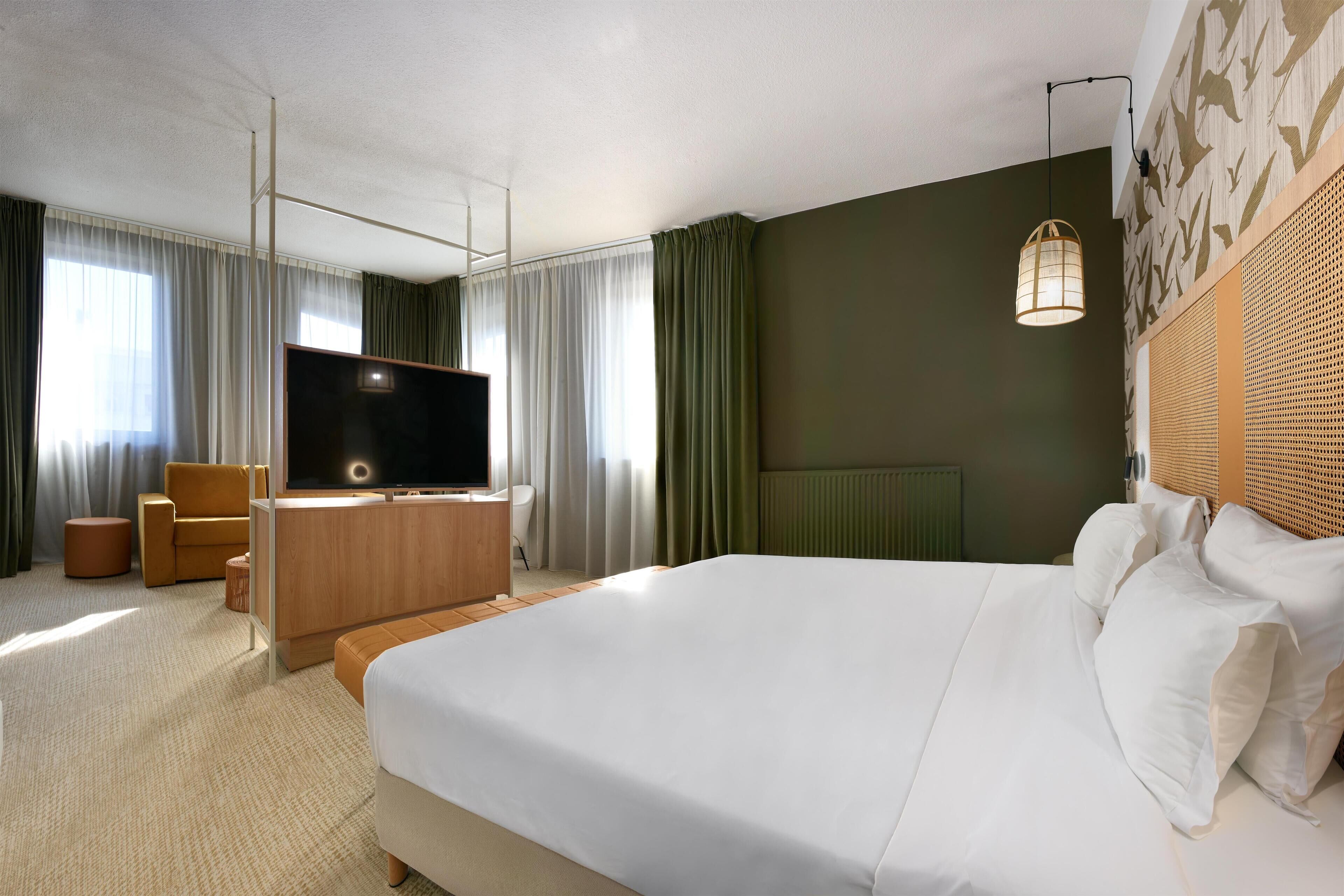 A modern hotel room with a large bed, a flat-screen TV, a wooden desk, and a comfortable armchair.