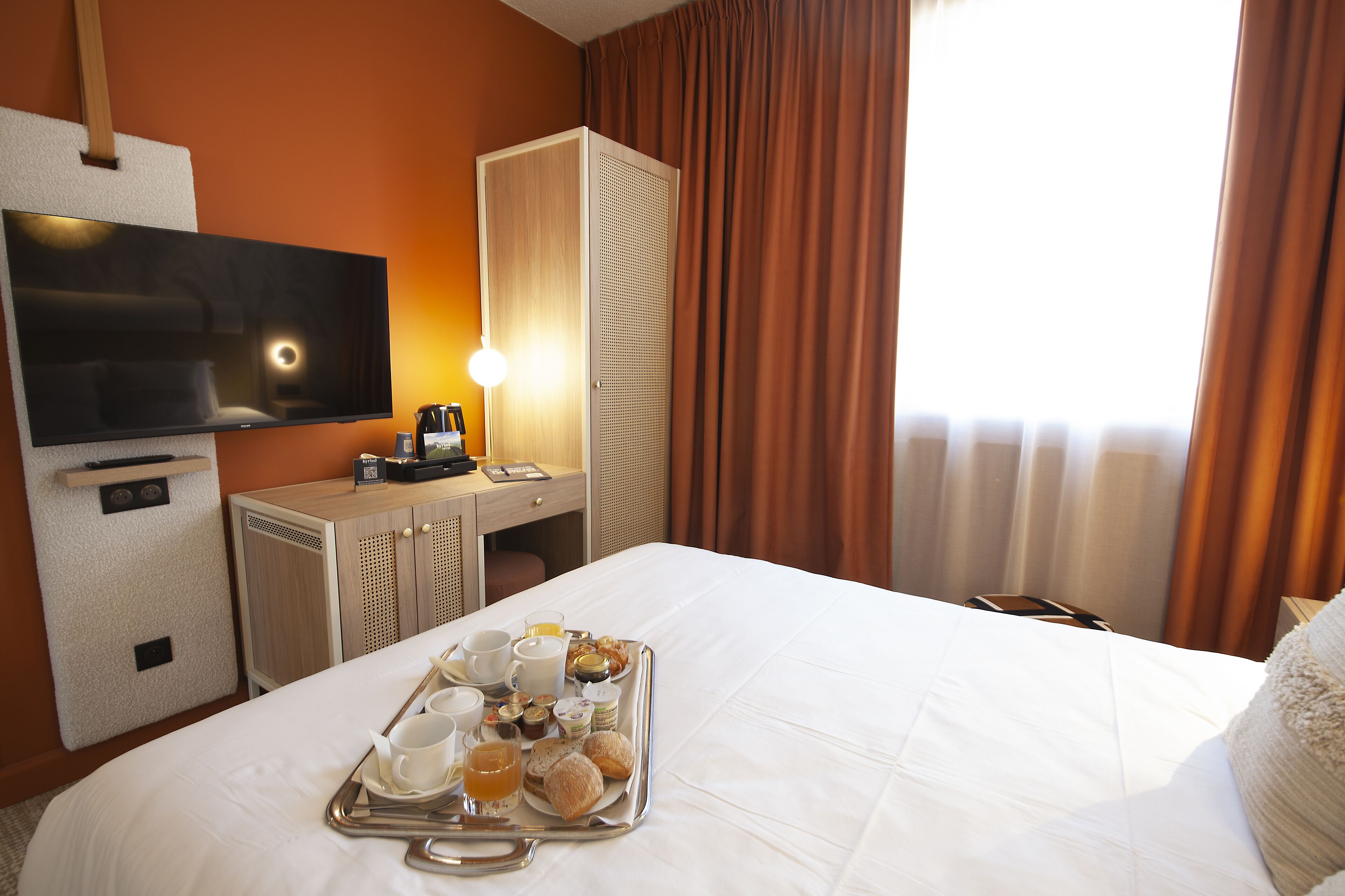 A hotel room with a bed, a TV, a desk with a lamp, and a tray with breakfast items.
