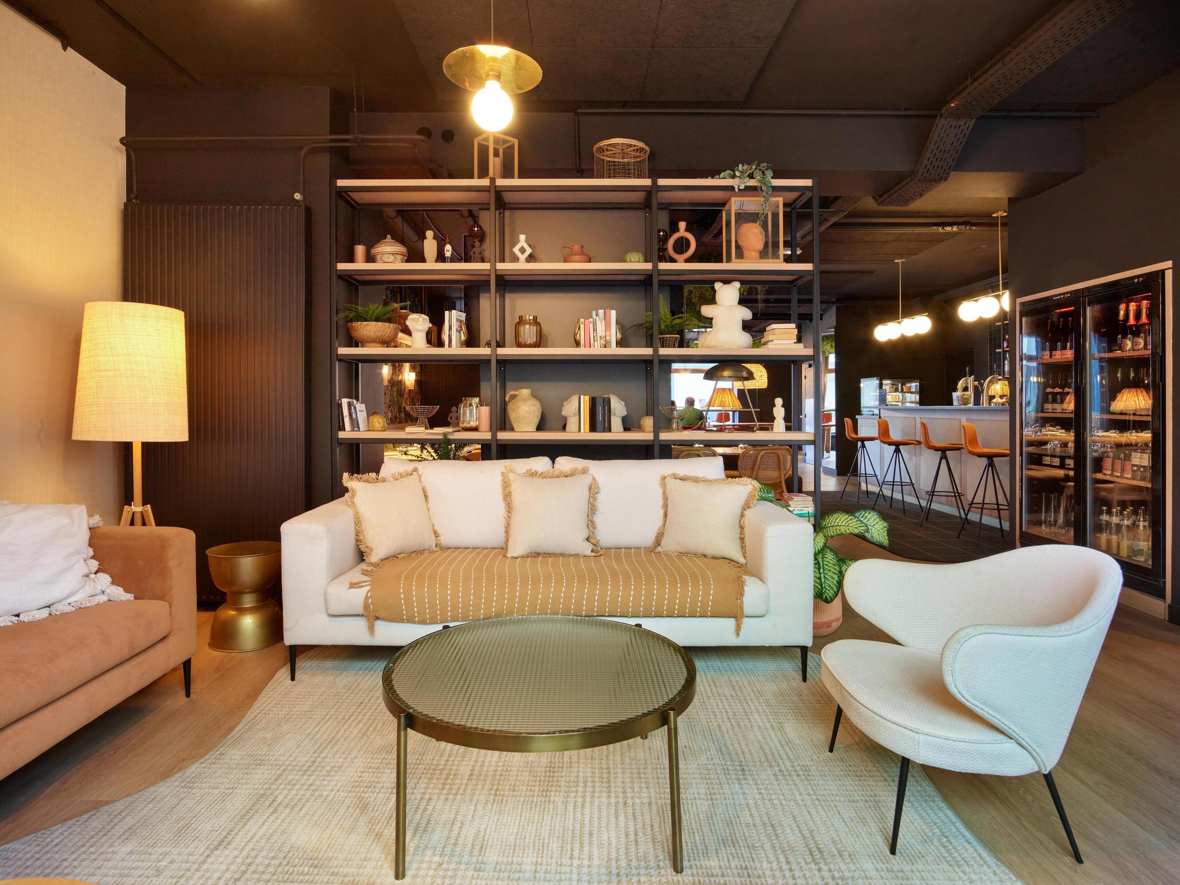Una sala moderna con un sofá, un sillón y una mesa de centro redonda. Hay una estantería llena de objetos decorativos y una zona de comedor con taburetes de bar.