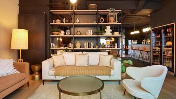 Una sala moderna con un sofá, un sillón y una mesa de centro redonda. Hay una estantería llena de objetos decorativos y una zona de comedor con taburetes de bar.