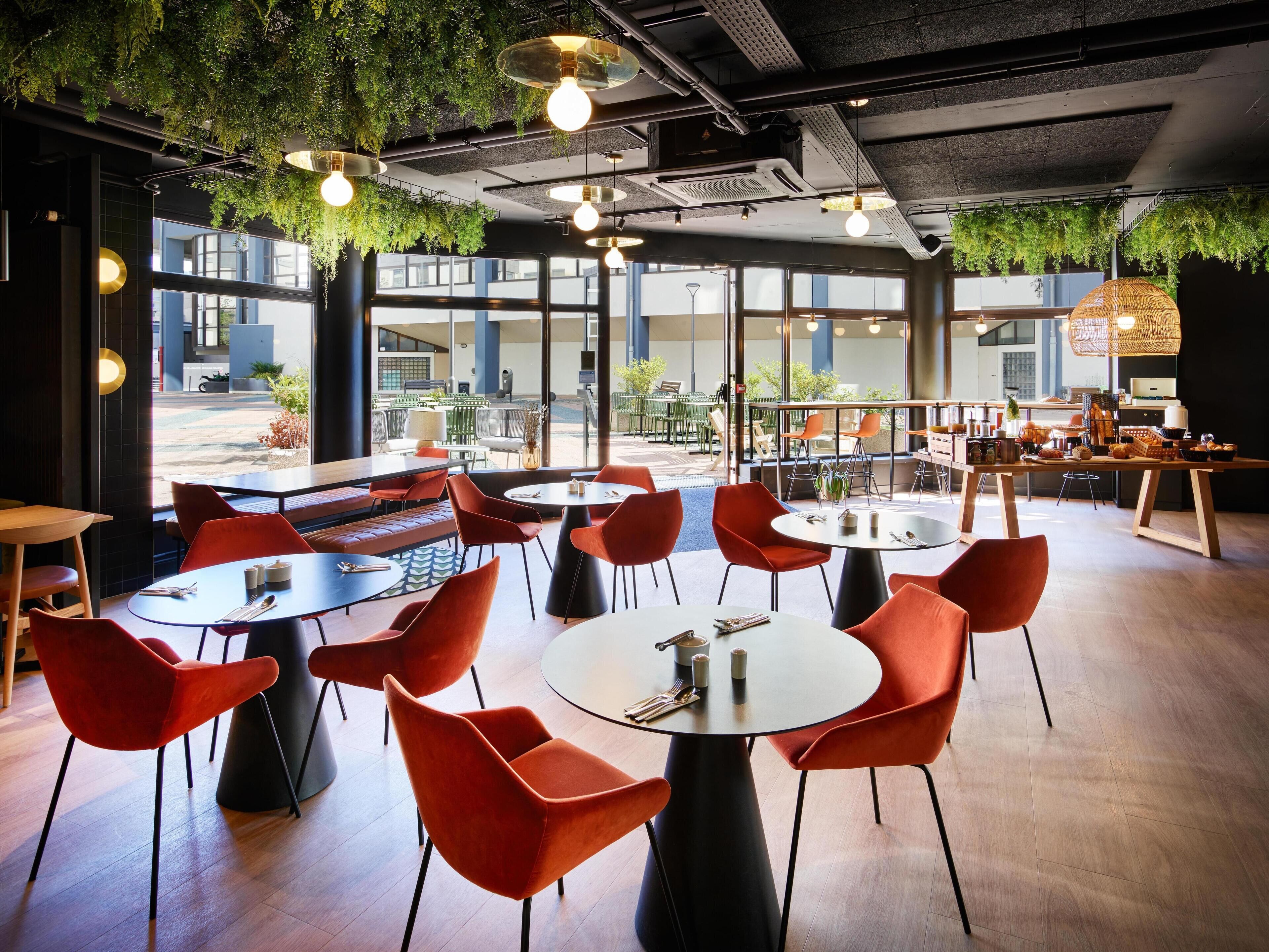 A modern cafe with round tables and red chairs, indoor plants, and large windows offering views of the street.