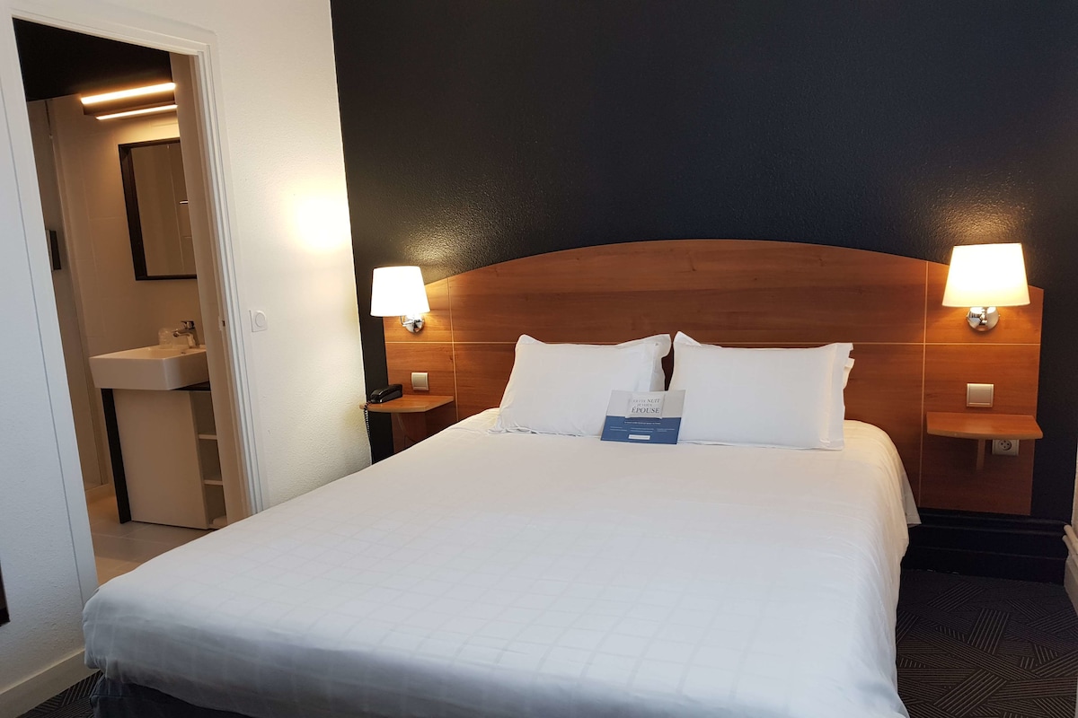 A hotel room with a large bed, two bedside lamps, a nightstand with a phone, and a small table with a sign.