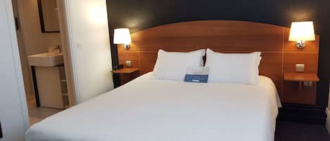 A hotel room with a large bed, two bedside lamps, a nightstand with a phone, and a small table with a sign.
