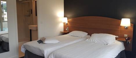 Habitación de hotel con dos camas, cabecera de madera, lámparas de noche, un espejo y un baño visible a través de una puerta abierta.