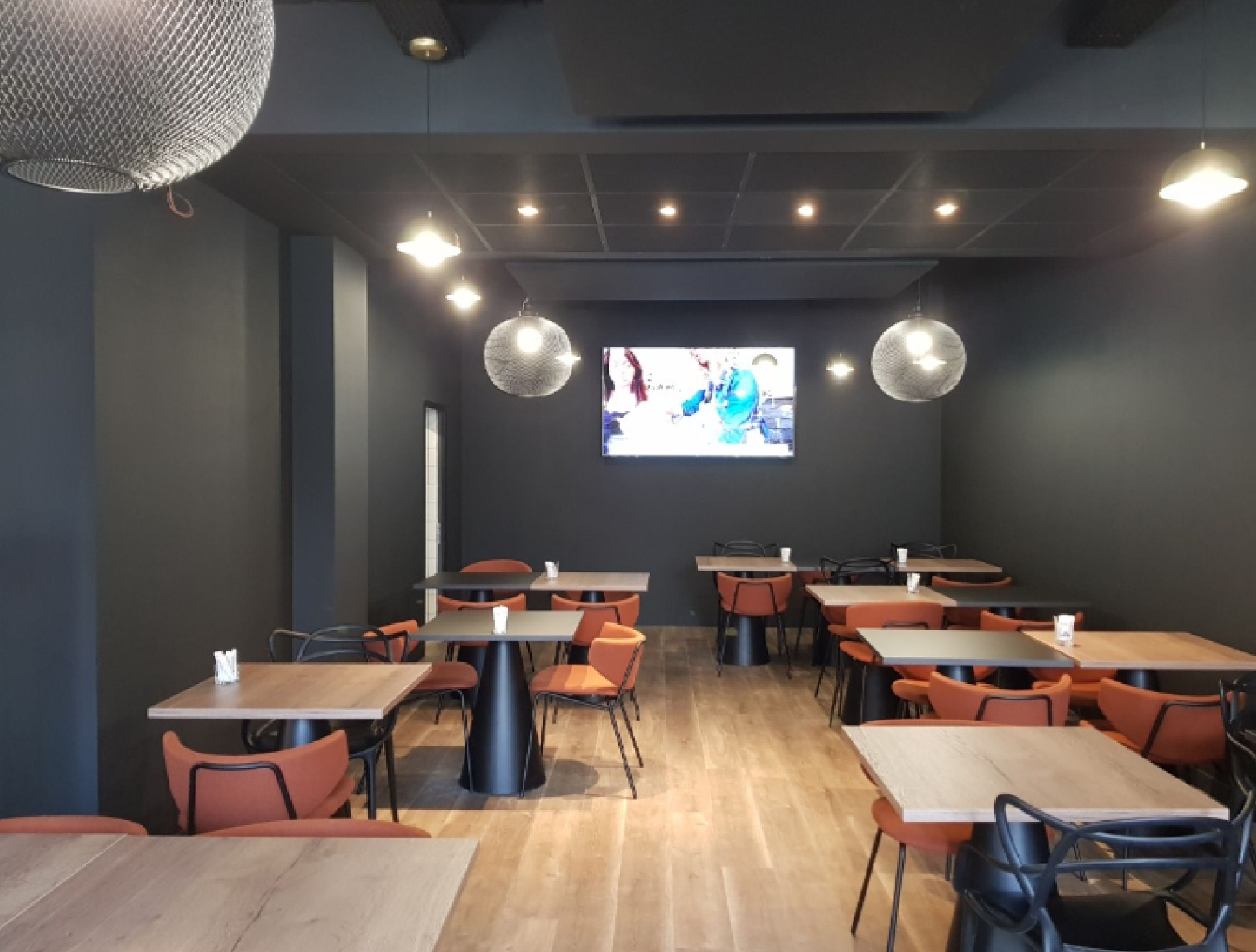 A modern restaurant interior with multiple tables and chairs, a large screen displaying a sports event, and pendant lighting.