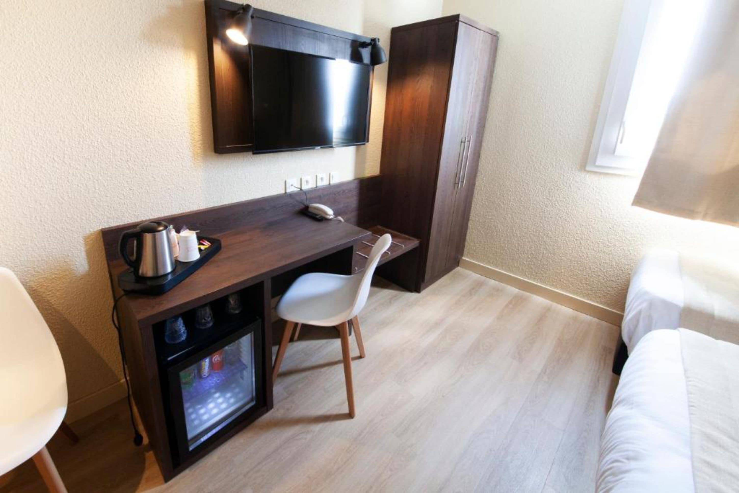Et lille hotelværelse med et træskrivebord, et tv ophængt over det, et minibar, en hvid stol og en seng med hvide sengetøj.