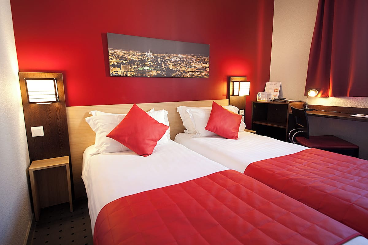 Une chambre d’hôtel avec deux lits, une literie rouge, une vue sur la ville, un bureau avec une lampe et une petite table de chevet.