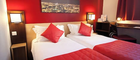 A hotel room with two beds, red bedding, a cityscape view, a desk with a lamp, and a small nightstand.