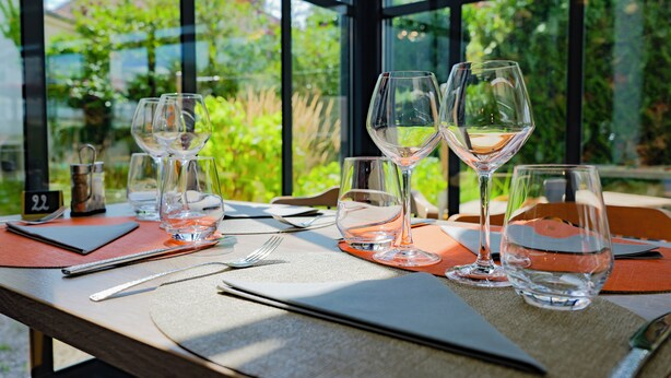 Une table dressée pour le dîner, avec des verres à vin et des couverts, un panneau indiquant le numéro 22, et une vue sur la verdure à travers de grandes fenêtres.