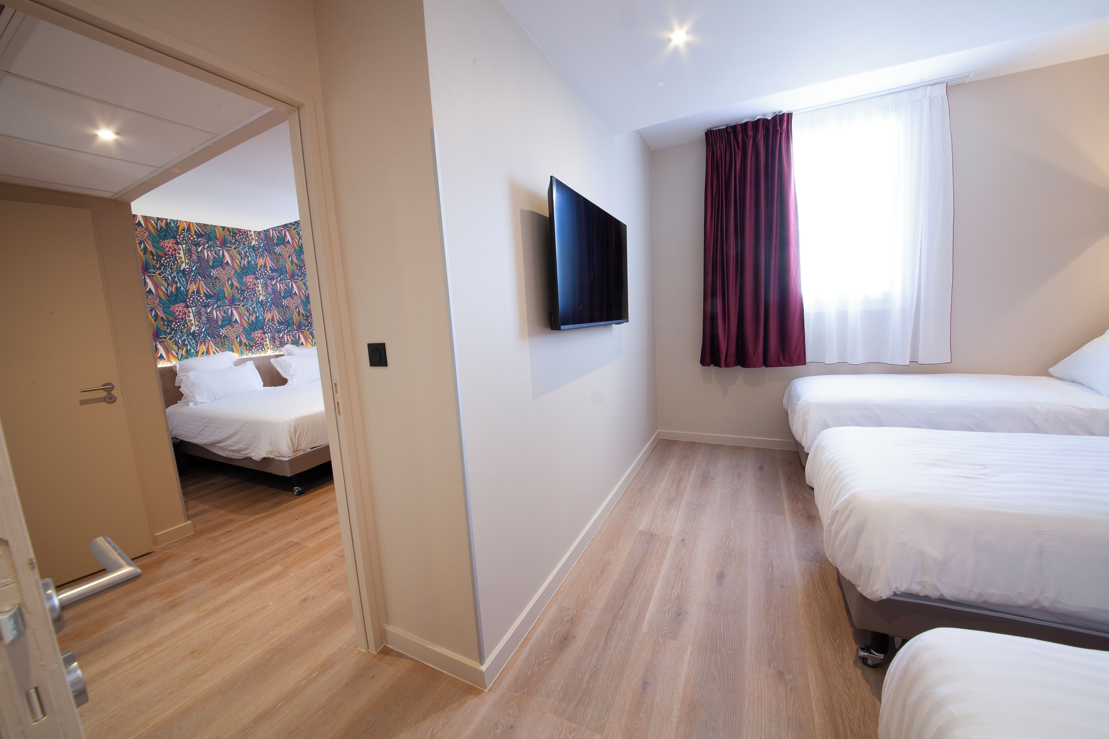 A hotel room with a wooden floor, a bed with white bedding, a television mounted on the wall, and a window with a curtain.