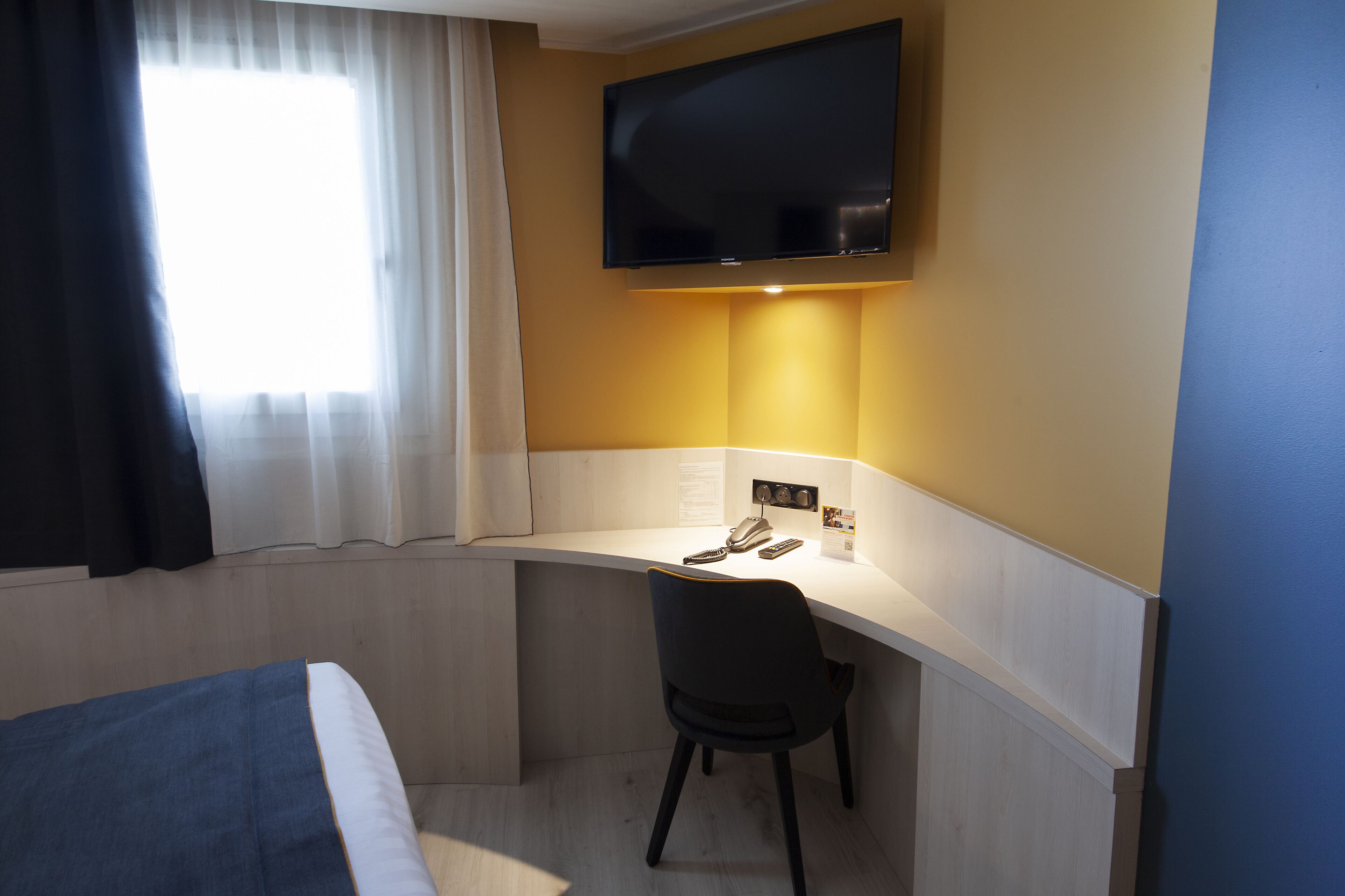 Habitación de hotel con cama, escritorio con silla, televisor de pantalla plana montado en la pared y ventana con cortinas.