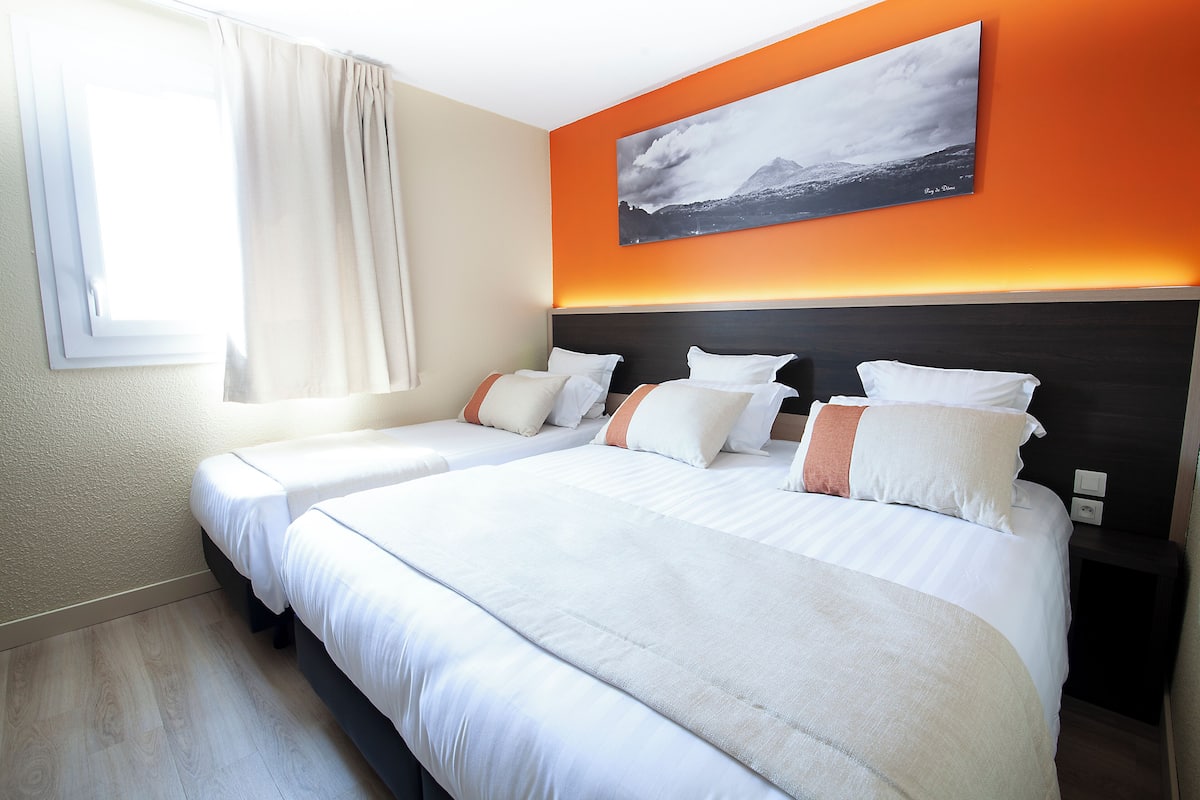 Une chambre d’hôtel avec deux lits, une grande fenêtre avec des rideaux, un mur d’accent orange et un tableau encadré au-dessus des lits.