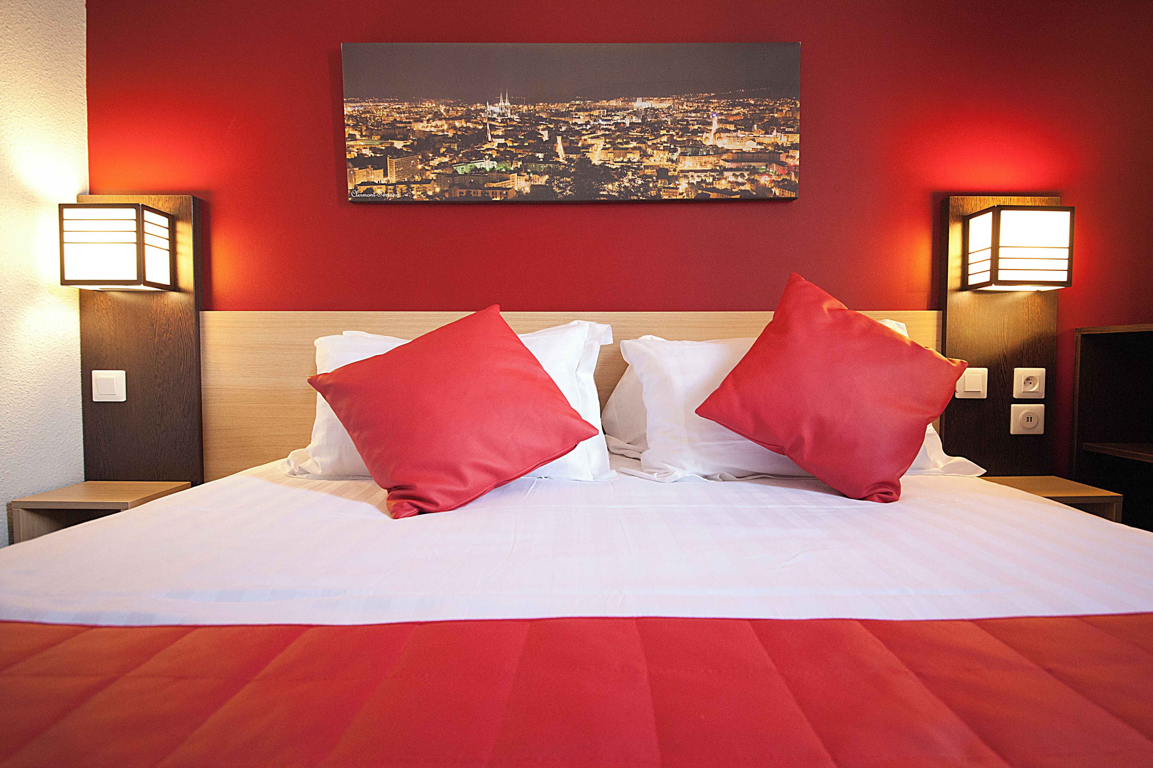 Una habitación de hotel con una cama grande, almohadas rojas y dos lámparas fijadas a la pared.
