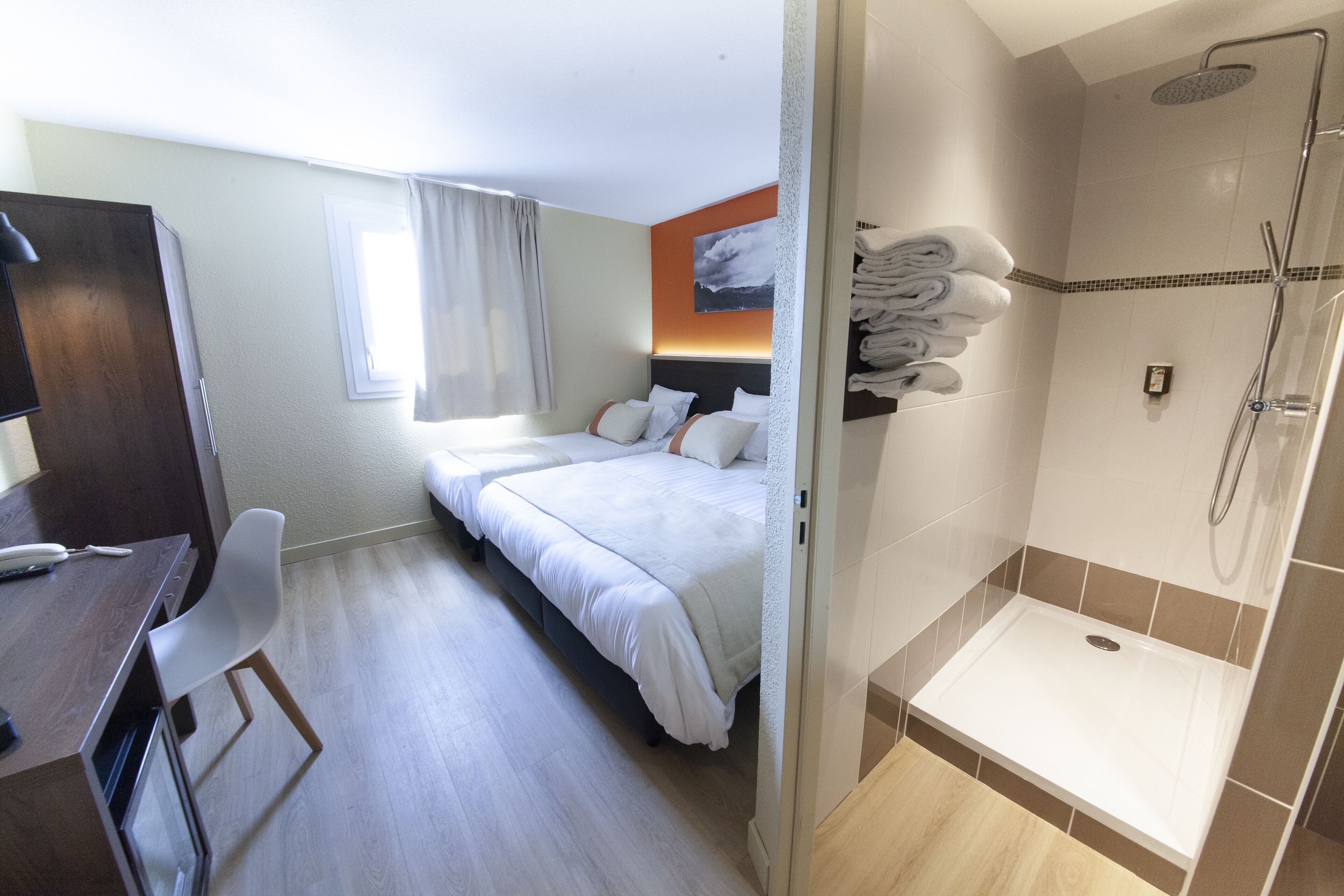 A hotel room with a bed, a desk, a chair, a shower area, and a window with curtains.