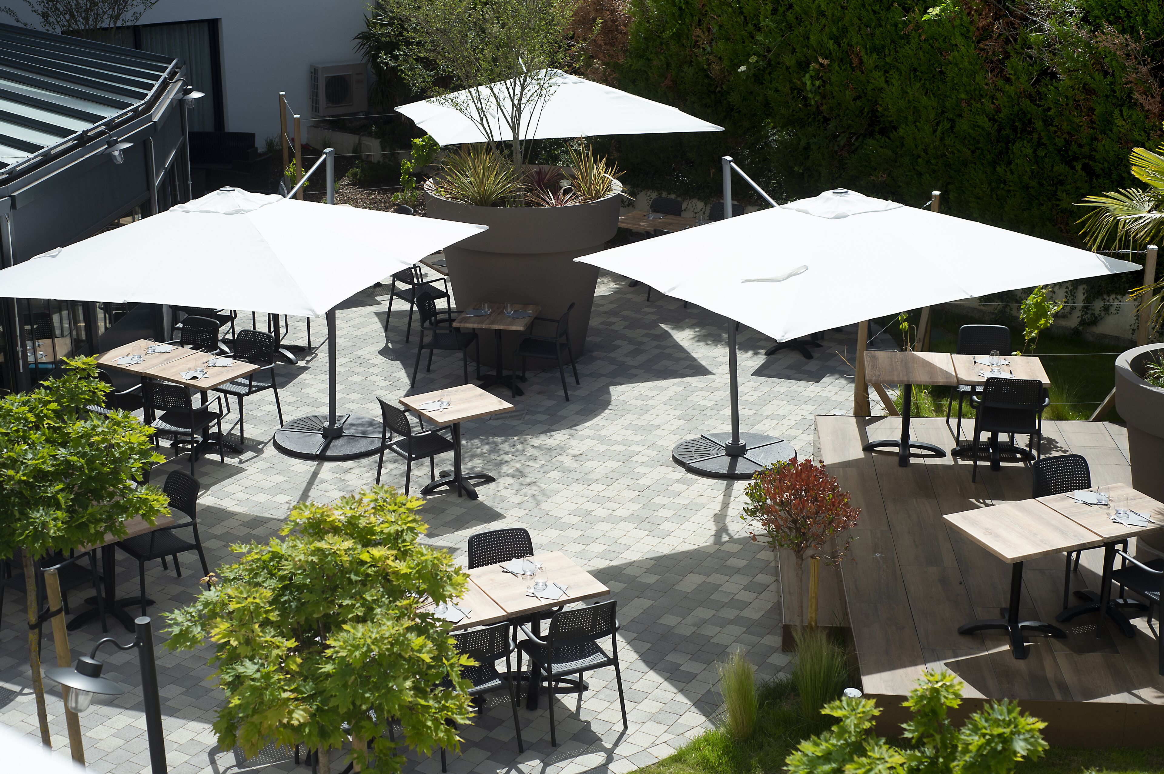 Un espace extérieur aménagé en terrasse, avec plusieurs tables et chaises, dont certaines sont placées sous de grands parasols blancs, le tout entouré de plantes en pot et d’arbres.