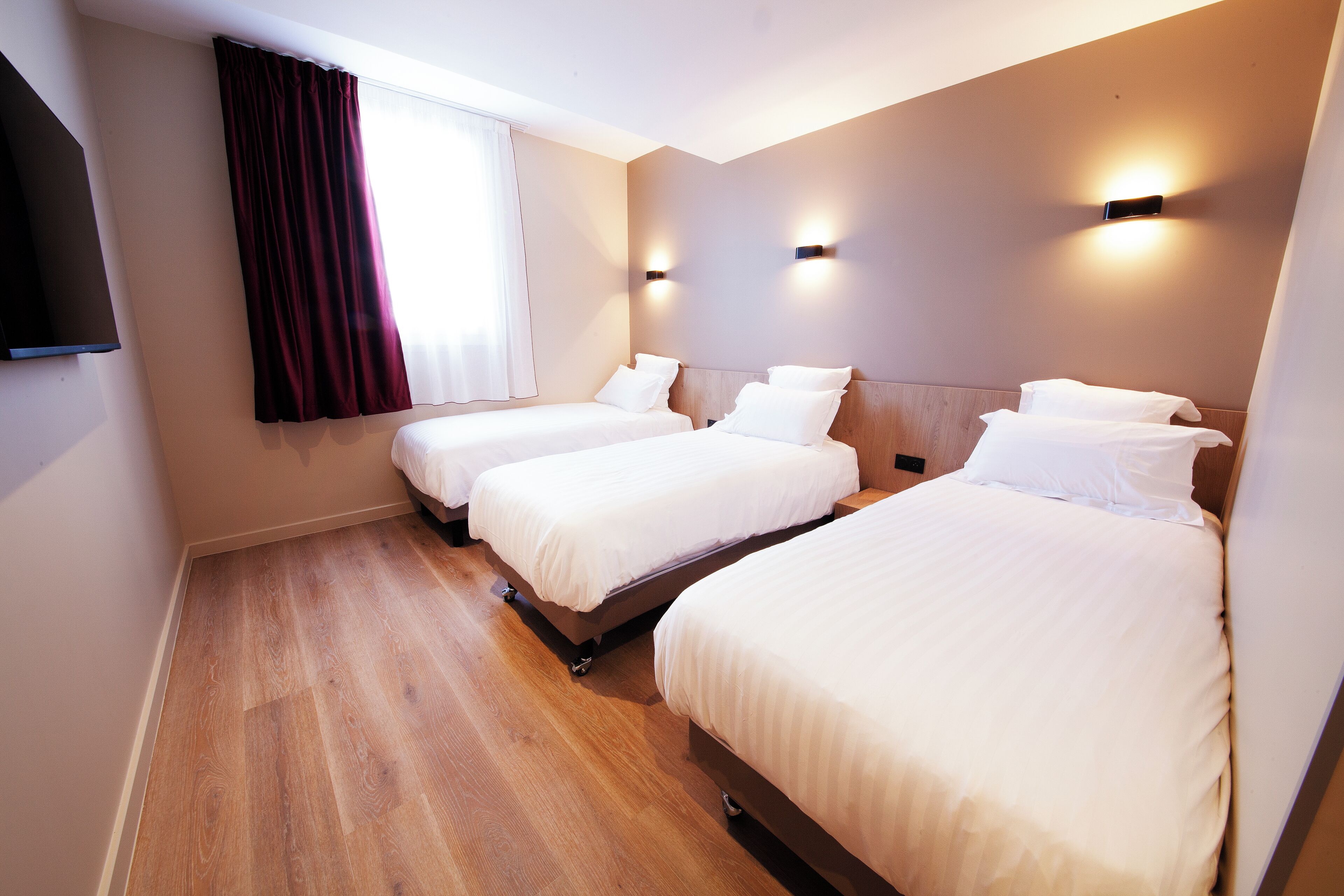 Habitación de hotel con tres camas individuales, piso de madera, un gran ventanal con cortinas e iluminación empotrada.