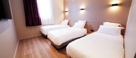 A hotel room with three single beds, a wooden floor, a large window with curtains, and wall-mounted lighting.