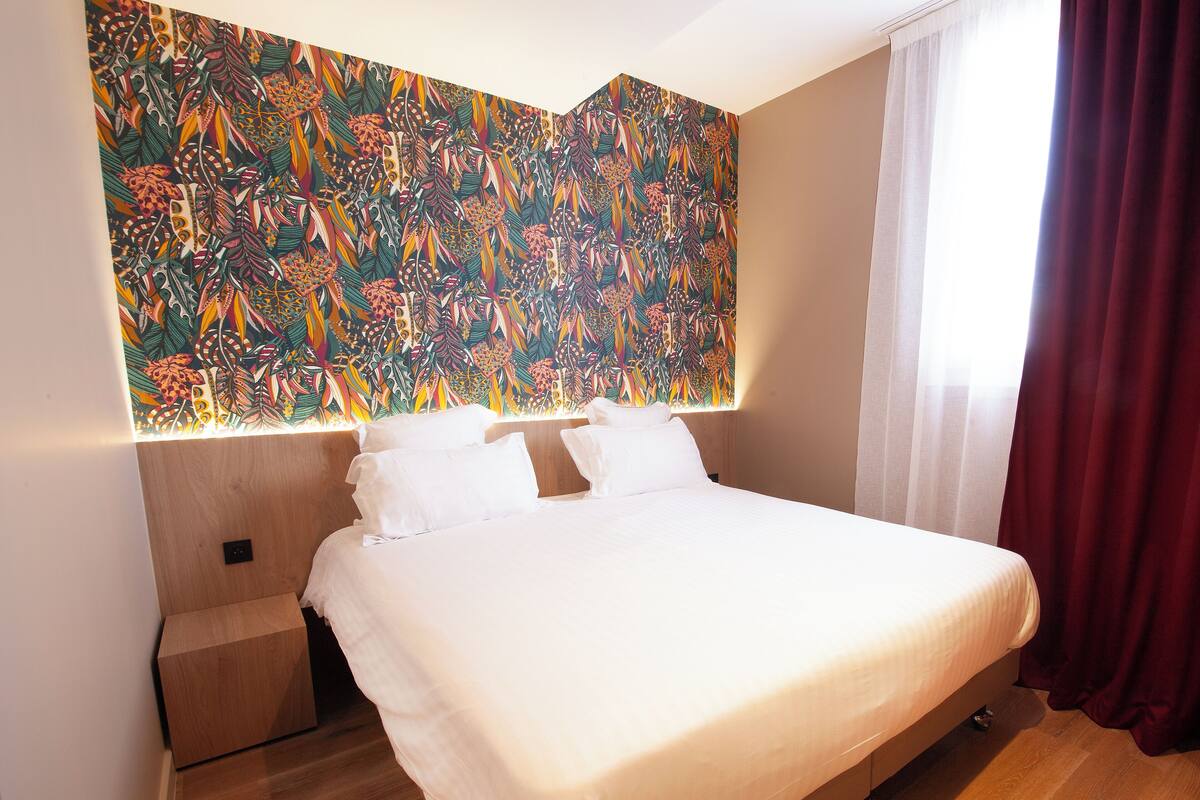 Une chambre d’hôtel moderne avec un grand lit, une tête de lit en bois et une fresque murale abstraite aux couleurs vives.