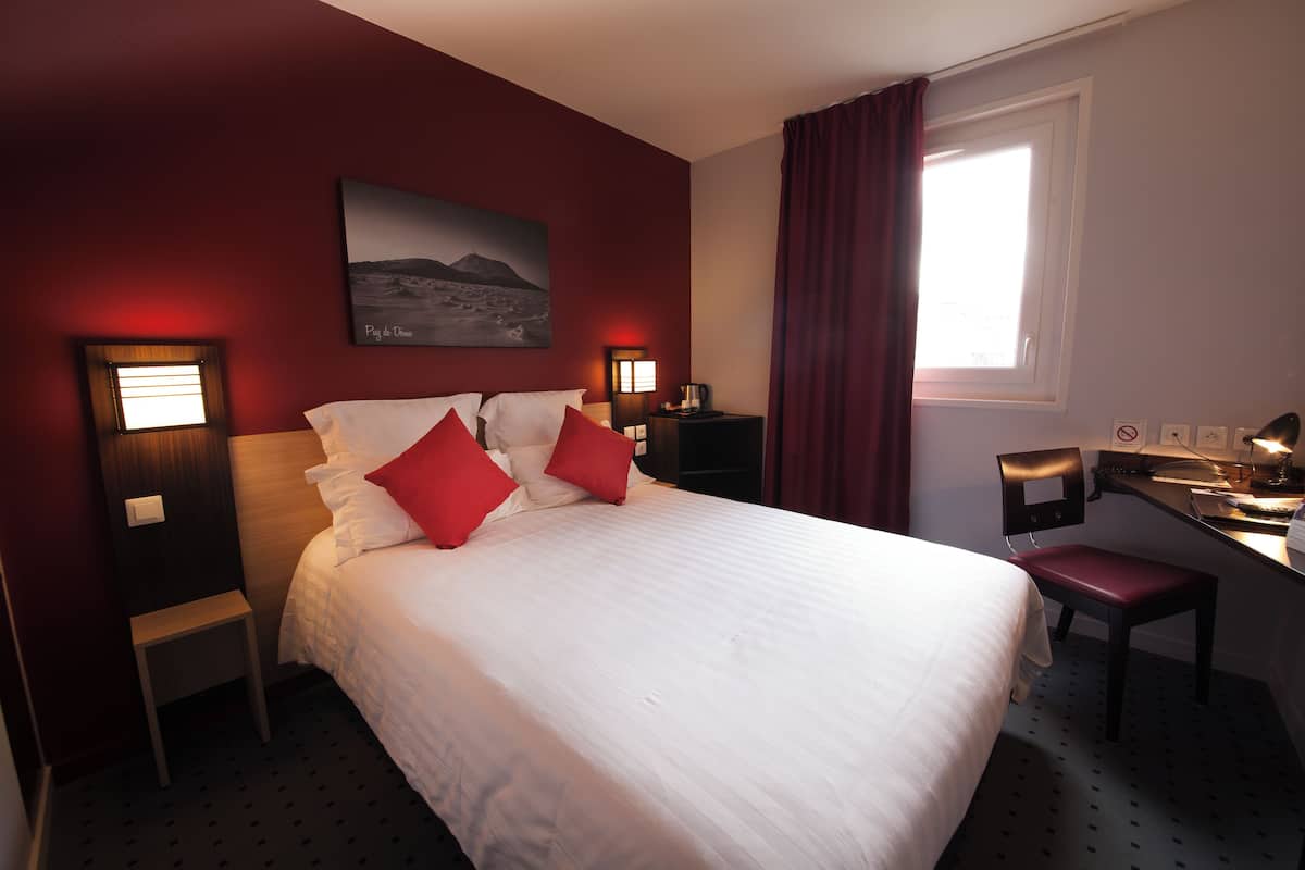 Une chambre d’hôtel avec un lit, un bureau, une chaise, une table de chevet, une lampe, un tableau et une fenêtre avec des rideaux.