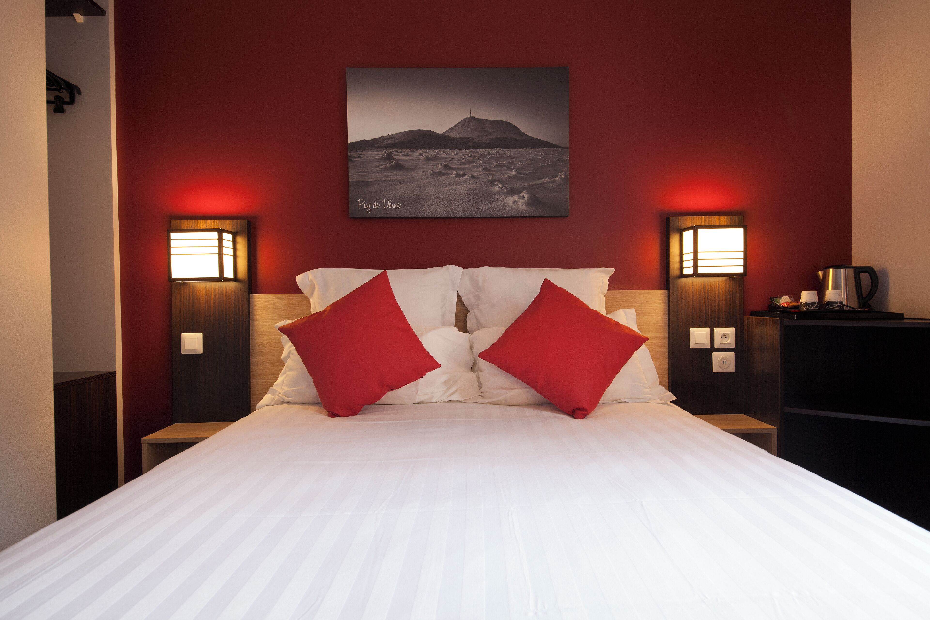 A hotel room with a large bed, red pillows, two bedside lamps, a painting of a mountain, and a nightstand with a coffee pot.