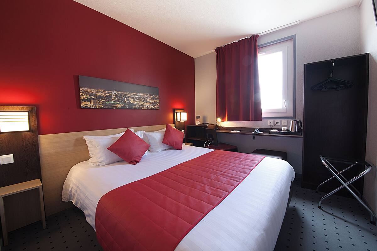 Une chambre d’hôtel avec un grand lit, des rideaux rouges, un tableau représentant un paysage urbain, un bureau avec une chaise et un porte-manteau.