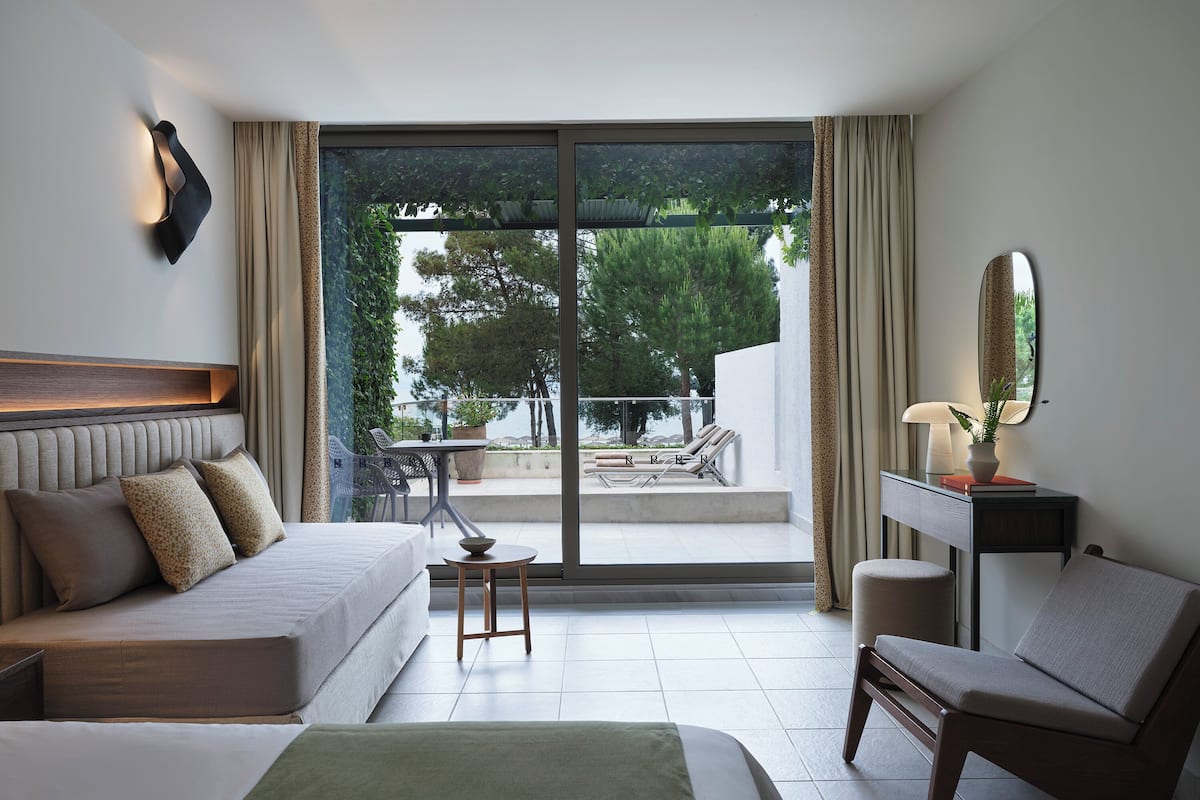 A modern hotel room with a bed, a desk, a chair, and a balcony with a view of trees and a pool.