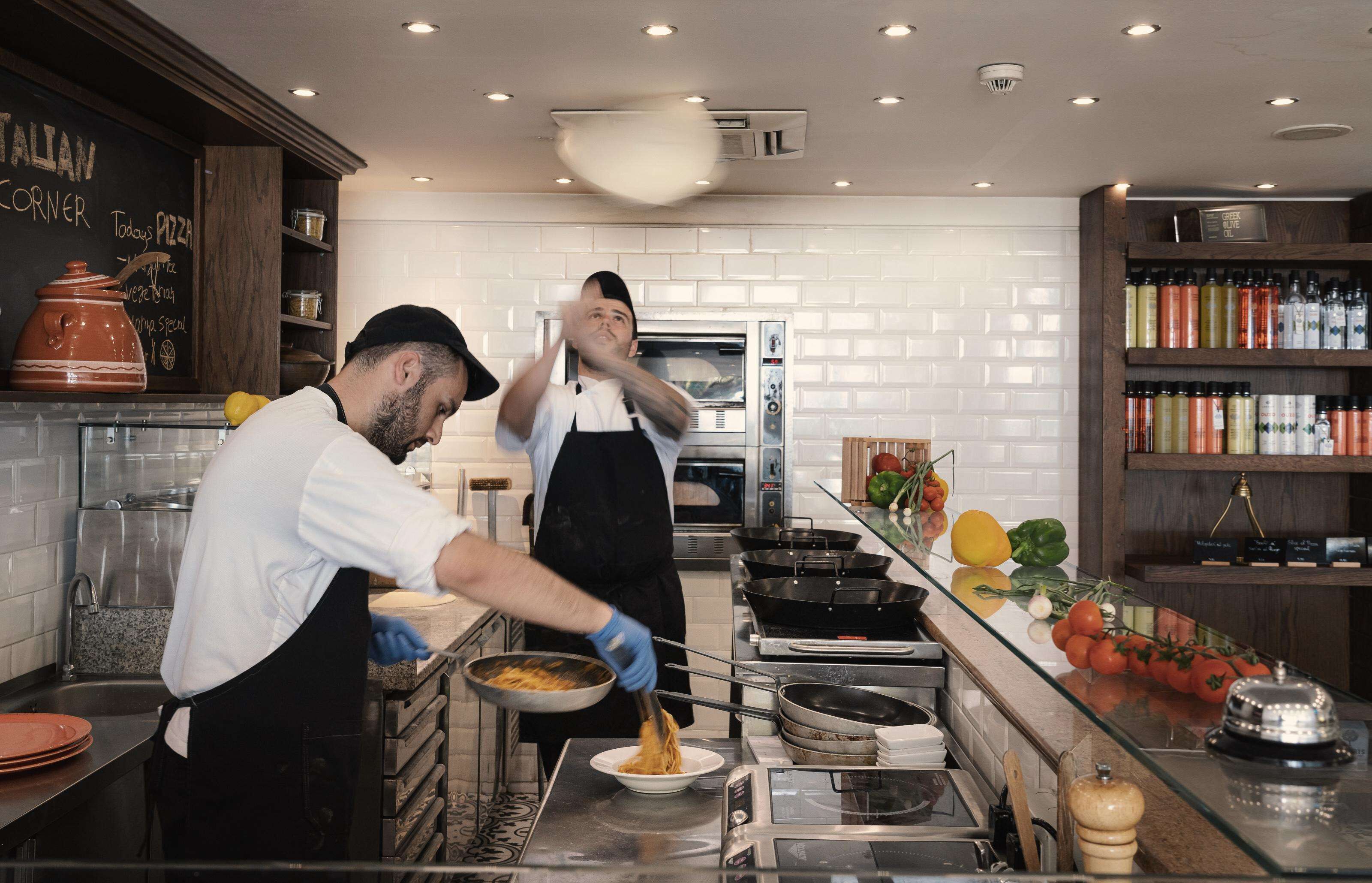 A kitchen with two chefs preparing food, a pizza oven, and various cooking utensils.