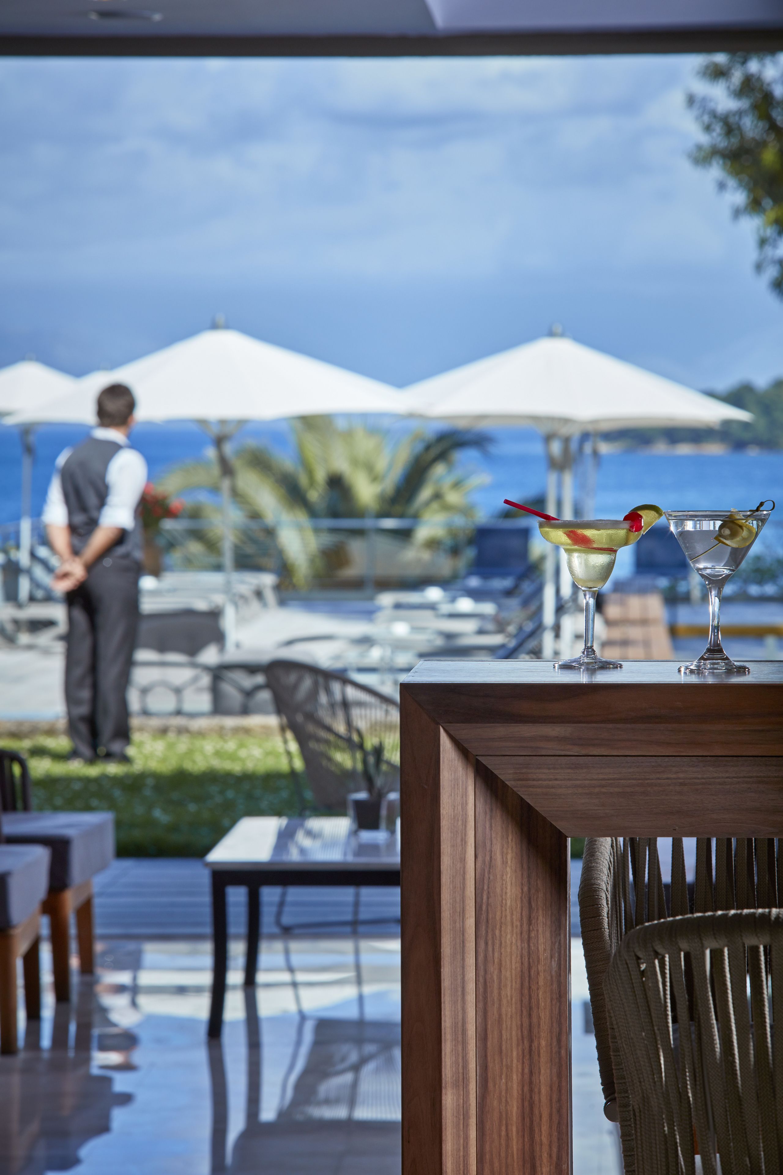 Uitzicht vanaf een balkon op een jachthaven, met twee parasols, een ober in de achtergrond en twee cocktails op een tafel.