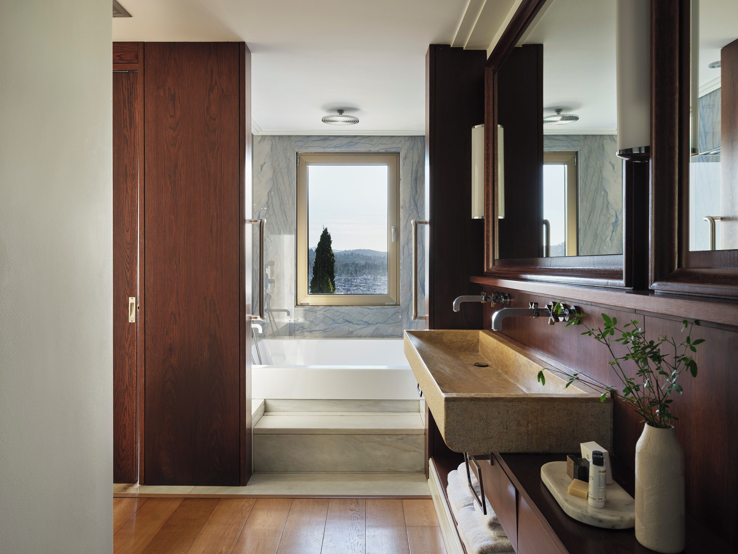 A modern bathroom with a wooden vanity, a large mirror, and a view of a cityscape through a window.